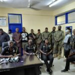 Police officers seated at a desk with microphones during a briefing, with soldiers and civilians standing in a room behind them.