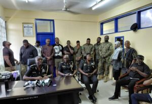 Police officers seated at a desk with microphones during a briefing, with soldiers and civilians standing in a room behind them.
