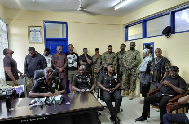 Police officers seated at a desk with microphones during a briefing, with soldiers and civilians standing in a room behind them.