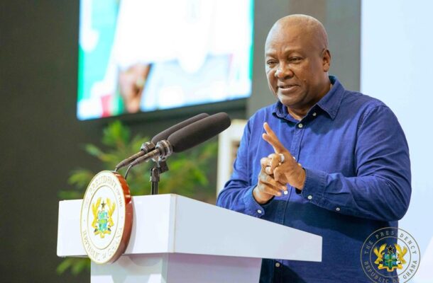 A man in a blue shirt speaks at a podium with two microphones, the presidential seal of Ghana visible on the lectern behind him.
