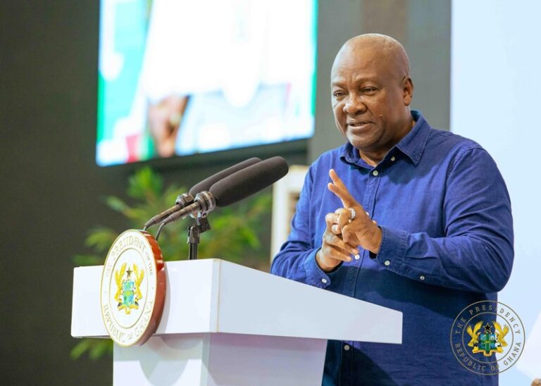 A man in a blue shirt speaks at a podium with two microphones, the presidential seal of Ghana visible on the lectern behind him.