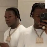 Twin mirror images of a man taking a selfie in a bathroom, wearing a white shirt and a large gold chain pendant.