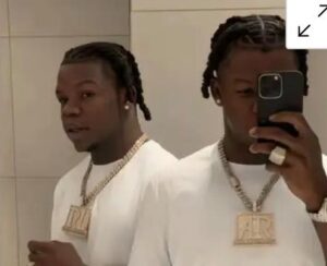 Twin mirror images of a man taking a selfie in a bathroom, wearing a white shirt and a large gold chain pendant.