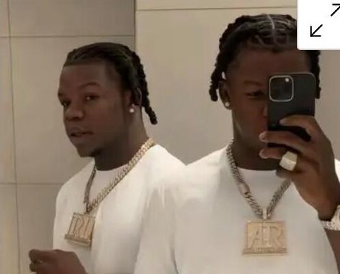 Twin mirror images of a man taking a selfie in a bathroom, wearing a white shirt and a large gold chain pendant.
