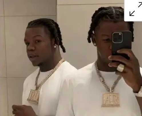 Twin mirror images of a man taking a selfie in a bathroom, wearing a white shirt and a large gold chain pendant.