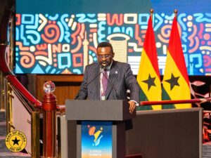 A man in a pinstripe suit speaks at a podium during a formal summit, with an abstract, colorful backdrop and Ghana and Senegal flags behind him.