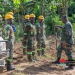 Ghana Armed Forces recruits in camouflage with yellow helmets receive a briefing from a supervising officer in a tropical outdoor training area beside a vehicle.