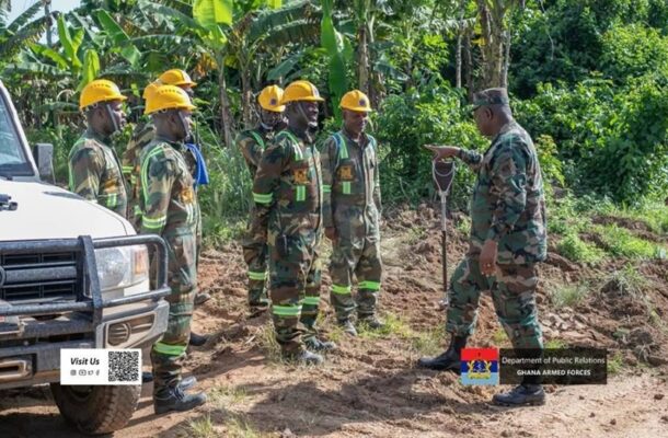 Ghana Armed Forces recruits in camouflage with yellow helmets receive a briefing from a supervising officer in a tropical outdoor training area beside a vehicle.