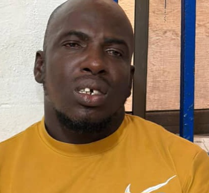 Portrait of a man with dark skin, wearing a mustard yellow shirt, showing missing teeth in the front.