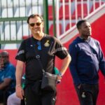 Two men stand near stadium seating; the man in front wears a black polo with a crest, sunglasses, and a crossbody bag, smiling at the camera.