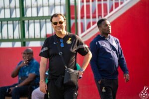 Two men stand near stadium seating; the man in front wears a black polo with a crest, sunglasses, and a crossbody bag, smiling at the camera.