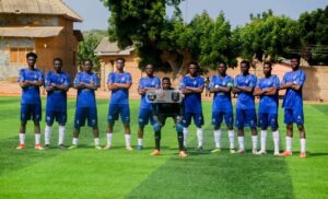 Soccer team in blue jerseys lined up on a green field, with the goalkeeper kneeling center and holding a banner.