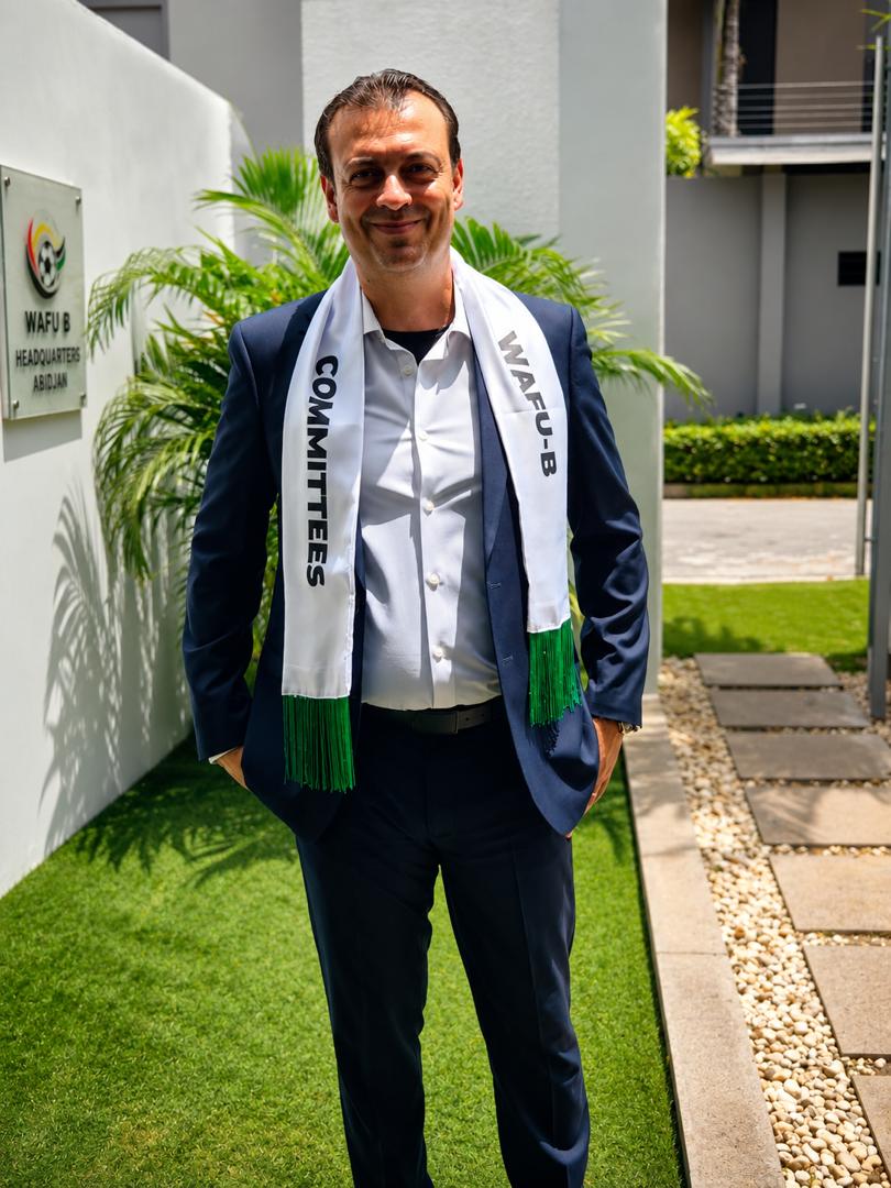 Smiling man in a navy suit wearing white scarves labeled 'COMMITTEES' and 'WAFU-B' standing outdoors near a modern building.