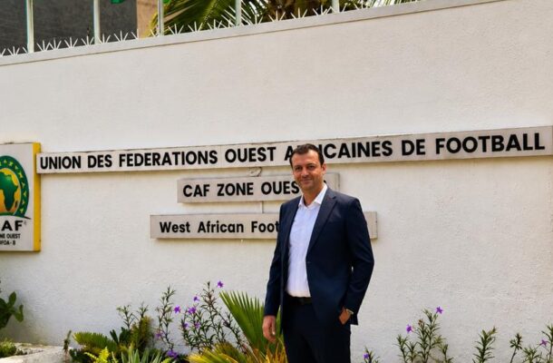 Man in a navy suit posing outside a West African football federation building under a row of flags and palm trees.