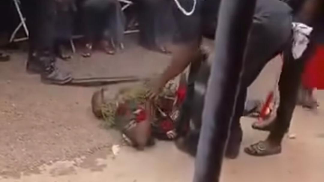Person in colorful clothing lying on a dusty street while a small crowd stands nearby.