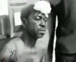 Man shirtless with a towel on his head, eyes closed, seated in a dim room.