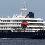 Large multi-deck cruise ship with dark blue hull and white superstructure sailing on calm open water, red lifeboat visible midship.
