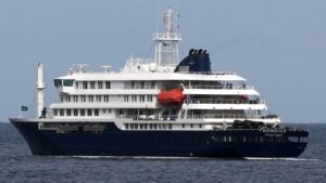 Large multi-deck cruise ship with dark blue hull and white superstructure sailing on calm open water, red lifeboat visible midship.