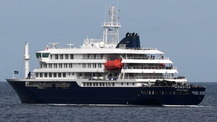 Large multi-deck cruise ship with dark blue hull and white superstructure sailing on calm open water, red lifeboat visible midship.