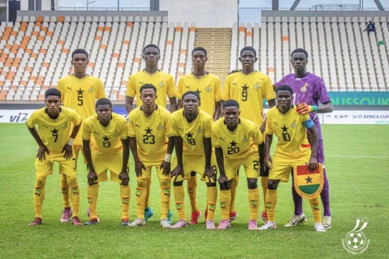 Youth soccer team in yellow uniforms poses for a group photo on a green field, with a purple-clad goalkeeper standing at the far right and players wearing numbered jerseys 5, 7, 13, 23, 18, 20, 10 in front row and 9, 6 in back row.