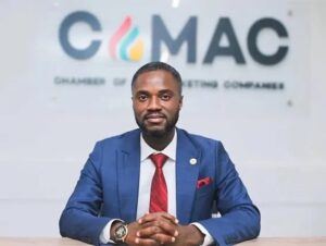 Portrait of a man in a blue suit and red tie seated at a desk with a CMAC sign behind him.