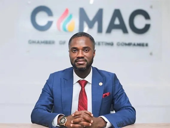Portrait of a man in a blue suit and red tie seated at a desk with a CMAC sign behind him.