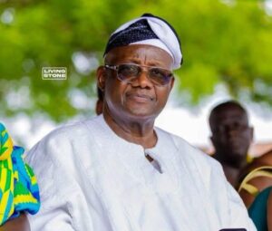 Older man in white traditional attire and head wrap, wearing glasses, at an outdoor event; Living Stone logo visible in background.