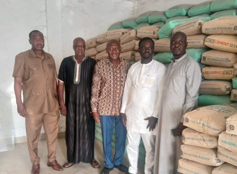 Five men stand side by side in a warehouse, with stacks of cement bags behind them.