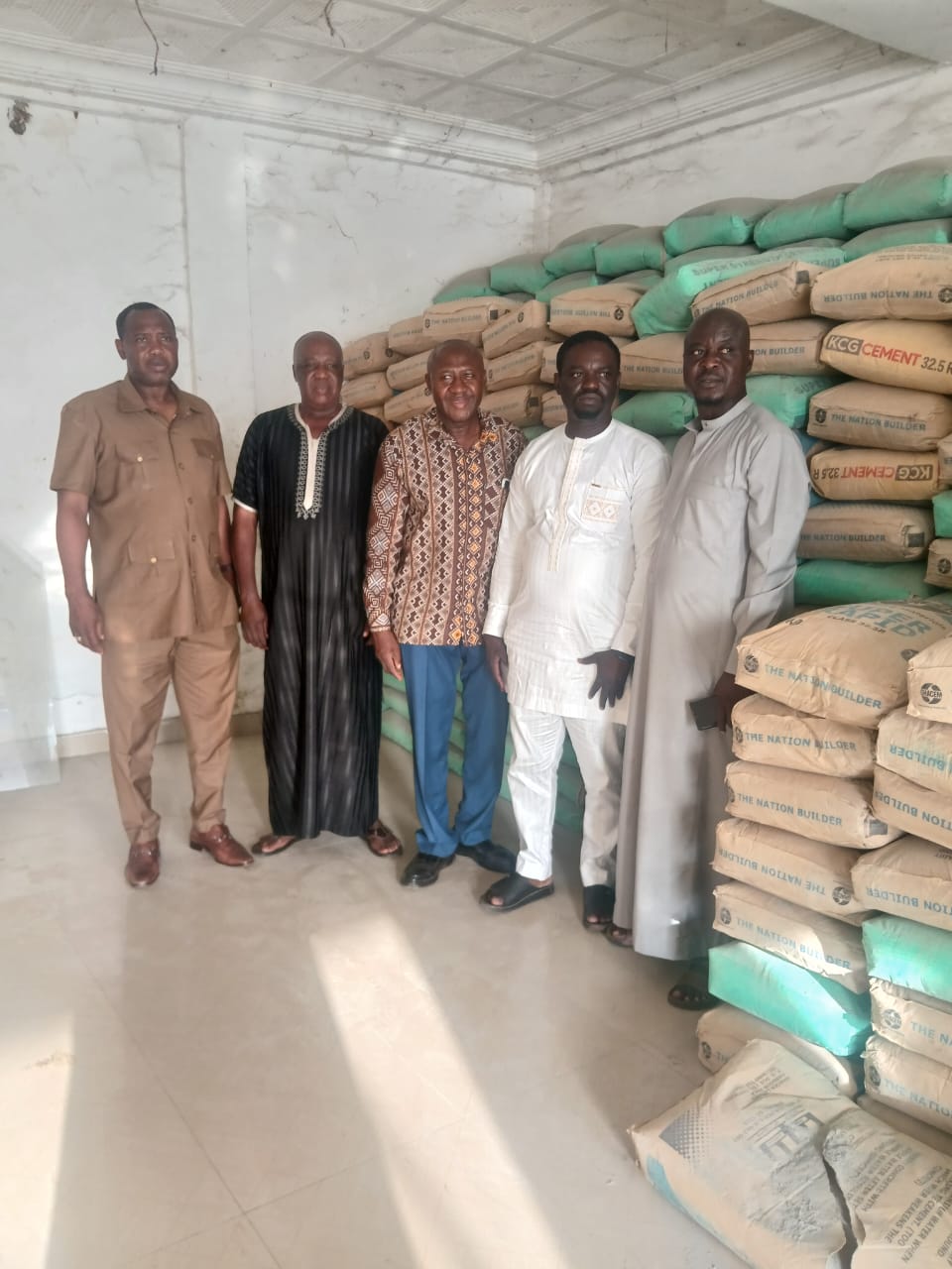IMG-20260517-WA0002 | Ghanaguardian.com Five men posing inside a room next to tall stacks of cement bags labeled 'The Nation Builder'.