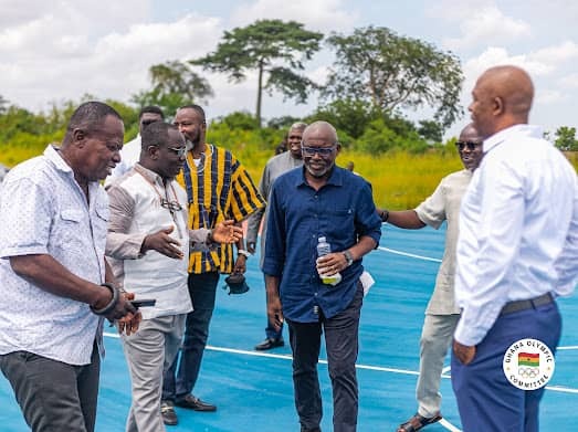 Group of men standing and talking on a blue outdoor court, one holding a bottle, in a rural setting with greenery in the background.