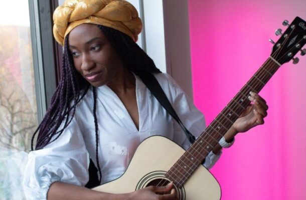 Woman with a yellow head wrap plays an acoustic guitar by a window, pink wall in the background.