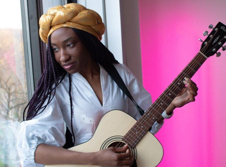 Woman with a yellow head wrap plays an acoustic guitar by a window, pink wall in the background.