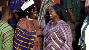 Two women in traditional striped garments smile and greet each other at a social gathering, with others in colorful outfits nearby.