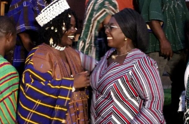 Two women in traditional striped garments smile and greet each other at a social gathering, with others in colorful outfits nearby.