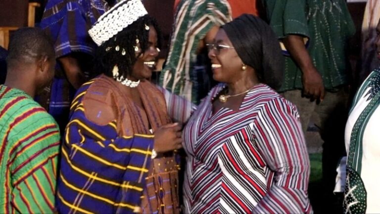 Two women in traditional striped garments smile and greet each other at a social gathering, with others in colorful outfits nearby.
