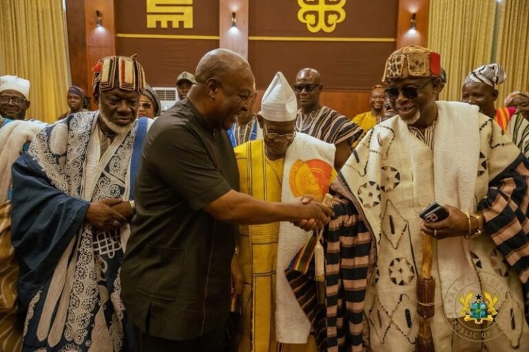 Dignitaries in traditional robes shake hands in a ceremonial room, with others watching in the background.