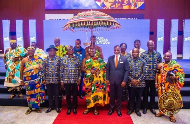 Group of people in colorful traditional African attire and a suit pose for a formal photo on a stage with a decorative umbrella during a business conference.