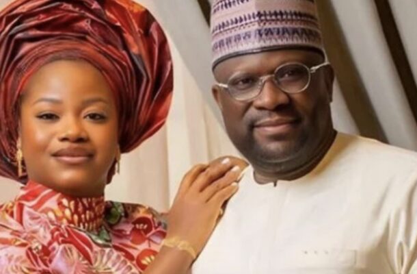 Couple in traditional Nigerian attire posing together; woman wears a red gele head wrap and floral dress, man in white agbada and cap.