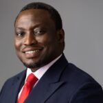 Portrait of a smiling Black businessman in a navy suit and red tie against a gray backdrop, looking at the camera.