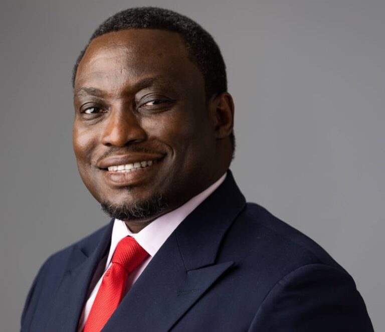 Portrait of a smiling Black businessman in a navy suit and red tie against a gray backdrop, looking at the camera.