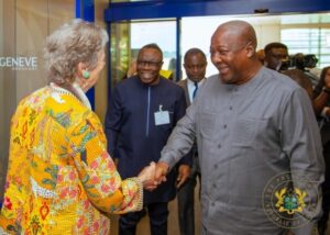 Two officials shake hands at a formal event, with a woman in a bright yellow patterned jacket on the left and a man in gray attire on the right, bystanders in the background.