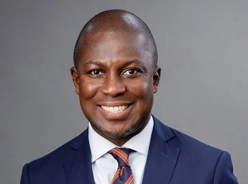 Professional headshot: smiling man in a navy suit and striped tie against a gray background.