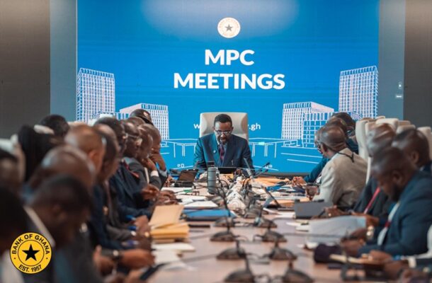 Officials in a boardroom around a long conference table under a blue backdrop reading 'MPC MEETINGS'.