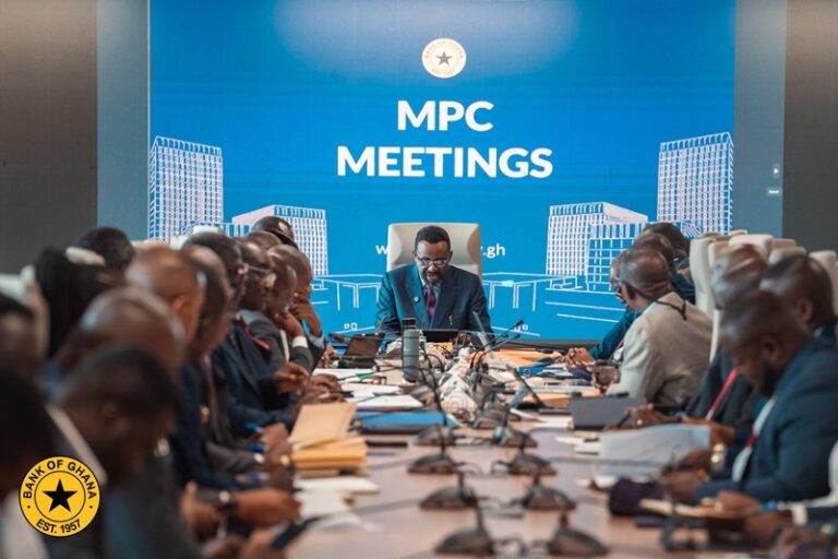 Officials in a boardroom around a long conference table under a blue backdrop reading 'MPC MEETINGS'.