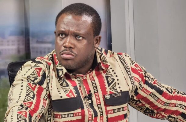 Man with a serious expression wearing a bold patterned shirt, seated indoors in a studio setting.
