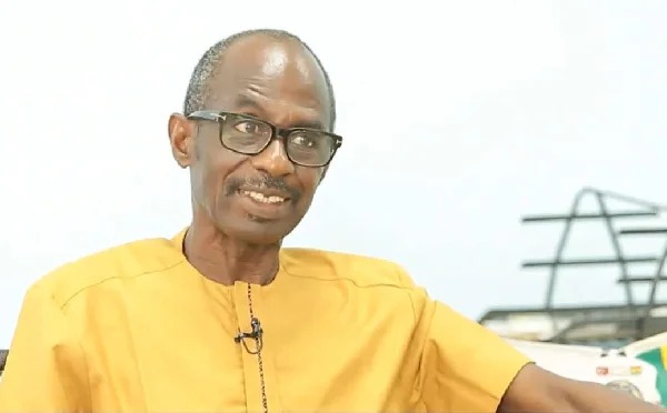 Smiling man wearing a yellow traditional shirt and glasses, seated and looking to the side.