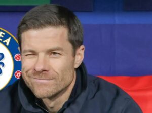 Close-up of a man with short dark hair and stubble winking at the camera, wearing a dark jacket, with blue and red backdrop and a partial logo to the left.