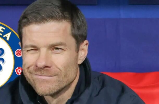 Close-up of a man with short dark hair and stubble winking at the camera, wearing a dark jacket, with blue and red backdrop and a partial logo to the left.
