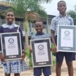 Three smiling students in uniforms stand outdoors holding framed certificates, celebrating achievements.
