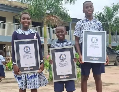 Three smiling students in uniforms stand outdoors holding framed certificates, celebrating achievements.